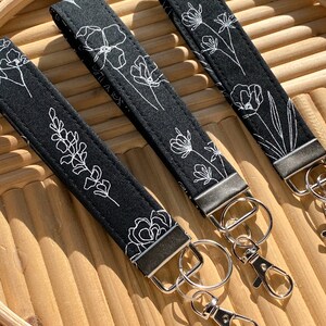 May include: Three black fabric wrist keychains with white floral line art designs. Each keychain has a silver-colored metal clasp and ring. The keychains are displayed on a light brown wooden tray.