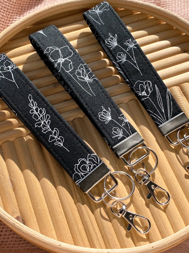 May include: Three black fabric wrist keychains with white floral line art designs. Each keychain has a silver-colored metal clasp and ring. The keychains are displayed on a light brown wooden tray.