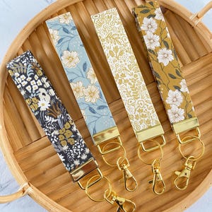 May include: Four floral patterned wrist keychains in navy, light blue, cream, and mustard yellow, each with a gold-tone metal clasp and ring. The keychains are arranged on a round, woven tray.