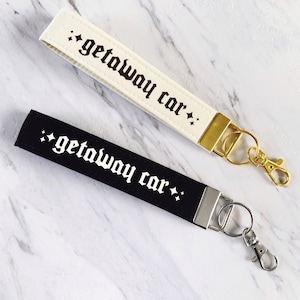 May include: Two wrist keychains, one cream and one black, with the phrase "getaway car" in gothic font. The cream keychain has gold hardware, while the black keychain has silver hardware. Both have a small star design.