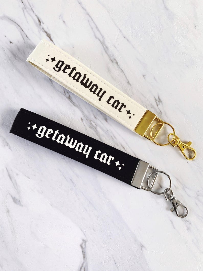 May include: Two wrist keychains, one cream and one black, with the phrase "getaway car" in gothic font. The cream keychain has gold hardware, while the black keychain has silver hardware. Both have a small star design.