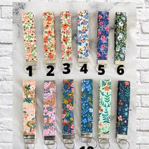 May include: Twelve fabric key fobs with floral patterns in various colors, including pink, orange, blue, and green. Each fob has a silver-colored metal ring and a loop for attaching keys. The text "CHOOSE YOUR FABRIC" is at the top.