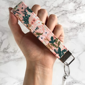 May include: A floral wristlet key fob with a pink background and green and white floral pattern. The key fob has a silver-colored metal clasp and a white tassel. The wristlet is held in a hand.