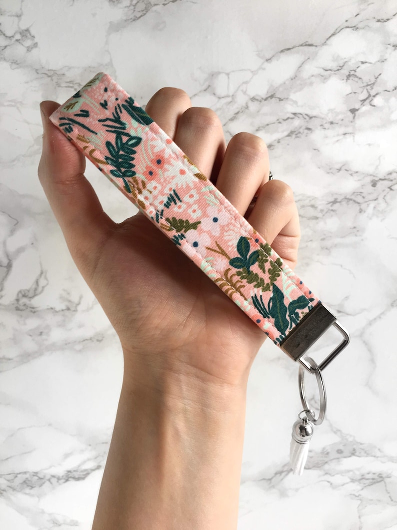 May include: A floral wristlet key fob with a pink background and green and white floral pattern. The key fob has a silver-colored metal clasp and a white tassel. The wristlet is held in a hand.