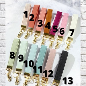 May include: A selection of 13 wrist keychains in various colors, including beige, coral, pink, and black. Each keychain has a gold-colored clasp and a fabric loop. The text "CHOOSE YOUR FABRIC" is at the top. The text "SEE COLOR DESCRIPTIONS IN ITEM DETAILS" is at the bottom.