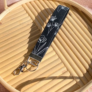 May include: Black wristlet key fob with a white floral design. The key fob has a silver-colored metal clasp and a matching metal piece at the top. The wristlet is lying on a light brown, woven tray.