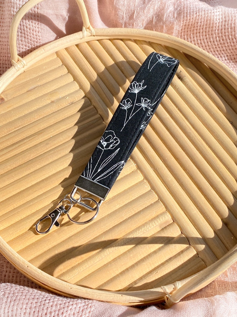 May include: Black wristlet key fob with a white floral design. The key fob has a silver-colored metal clasp and a matching metal piece at the top. The wristlet is lying on a light brown, woven tray.