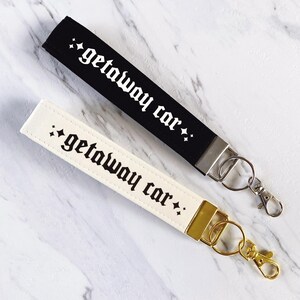 May include: Two keychains, one black and one cream-colored, each with the phrase "getaway car" in a gothic font. The black keychain has a silver clasp, while the cream-colored one has a gold clasp. Both are on a marble surface.