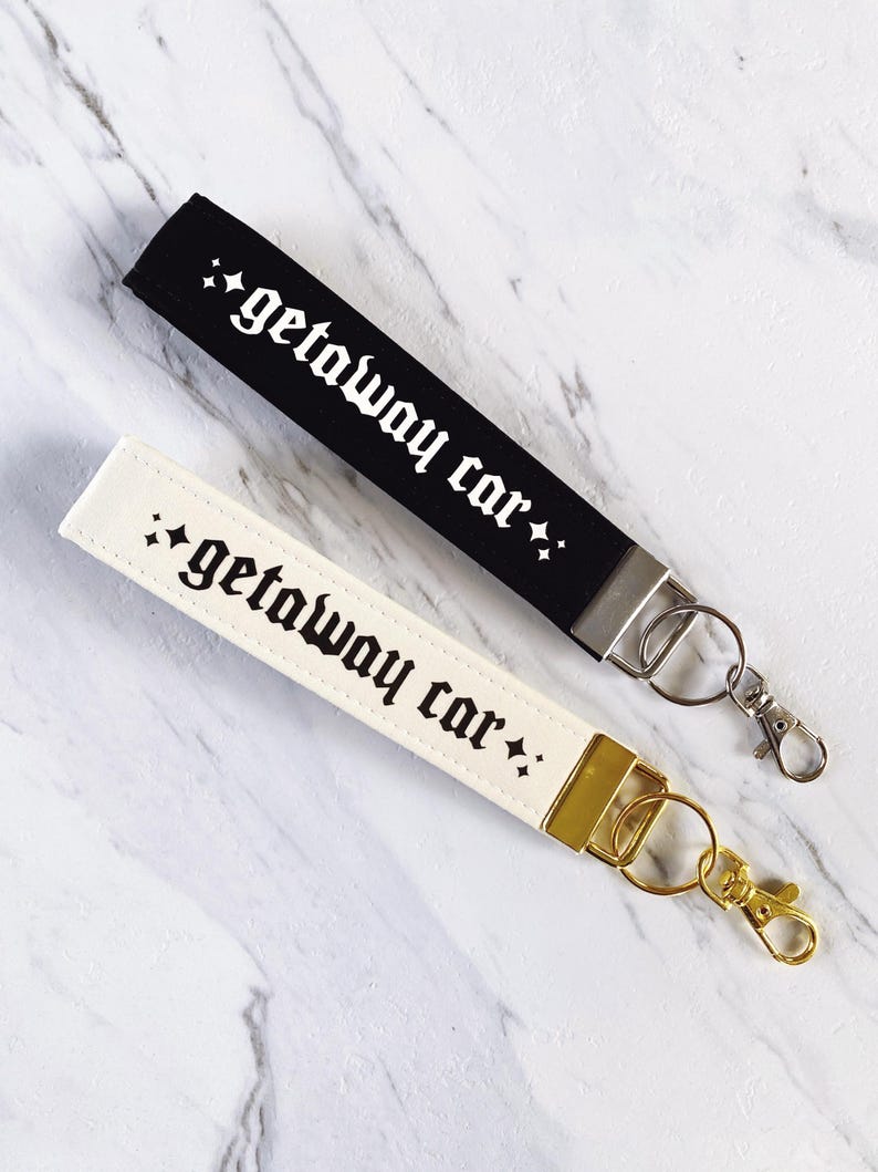 May include: Two keychains, one black and one cream-colored, each with the phrase "getaway car" in a gothic font. The black keychain has a silver clasp, while the cream-colored one has a gold clasp. Both are on a marble surface.