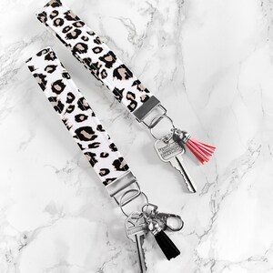 May include: Two key wristlets with a white background and a leopard print pattern in black and tan. Each wristlet has a silver metal clasp and key ring. One has a pink tassel, the other a black tassel.