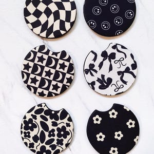 May include: Six black and white car coasters with various patterns. Designs include a checkerboard, smiley faces, stars and moons, bows, flowers, and daisy patterns. Each coaster has a curved cutout for easy placement and removal from a car cup holder.