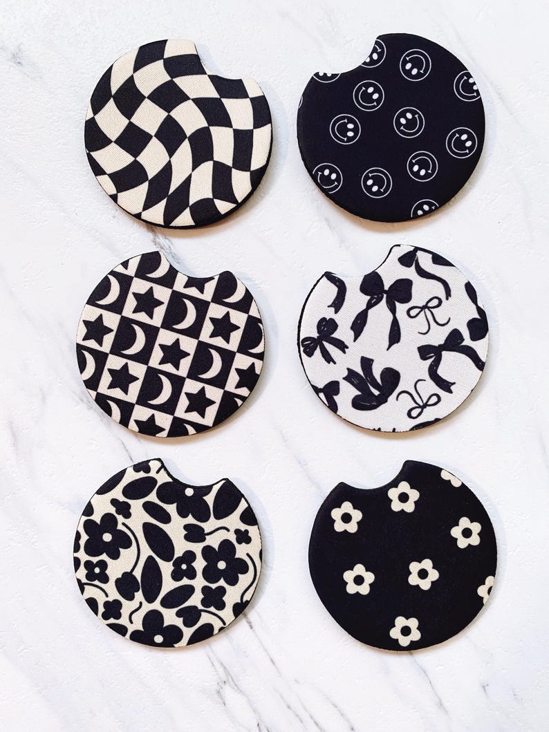 May include: Six black and white car coasters with various patterns. Designs include a checkerboard, smiley faces, stars and moons, bows, flowers, and daisy patterns. Each coaster has a curved cutout for easy placement and removal from a car cup holder.