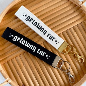 May include: Two wrist keychains, one white and one black, each with the text "getaway car" in a gothic font. The white keychain has a gold clasp, and the black keychain has a silver clasp. Both are on a woven tray.