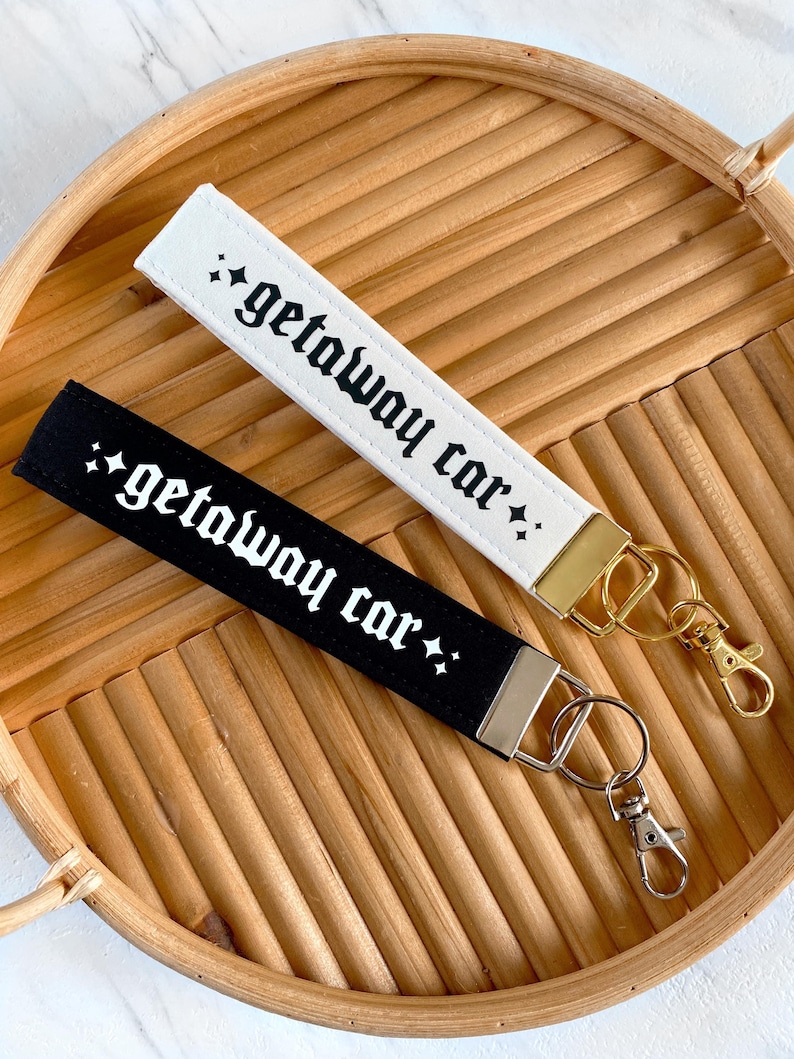 May include: Two wrist keychains, one white and one black, each with the text "getaway car" in a gothic font. The white keychain has a gold clasp, and the black keychain has a silver clasp. Both are on a woven tray.