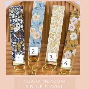 May include: Four fabric key fobs with gold hardware, each featuring a different floral pattern. The patterns include dark meadow, blue floral, ditsy gold, and gold floral designs. The text "CHOOSE YOUR FABRIC" is at the top.