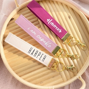 May include: Three wrist keychains in shades of pink, purple, and cream, each with gold-colored hardware. The keychains are personalized with the names "Harper" and "Lauren", and the phrase "I am capable" in white lettering. They rest on a woven tray.