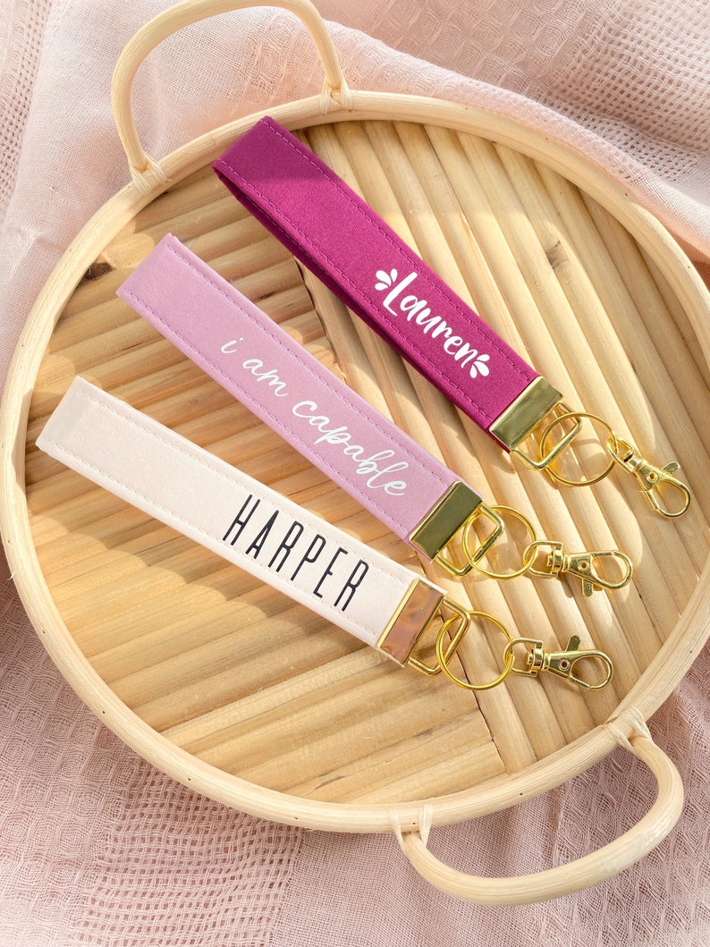 May include: Three wrist keychains in shades of pink, purple, and cream, each with gold-colored hardware. The keychains are personalized with the names "Harper" and "Lauren", and the phrase "I am capable" in white lettering. They rest on a woven tray.