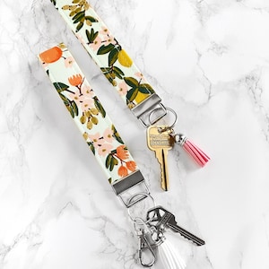 May include: Two floral print wrist keychains with silver hardware. One keychain has a pink tassel, the other a white tassel. The fabric features a light green background with orange, pink, and yellow floral designs.
