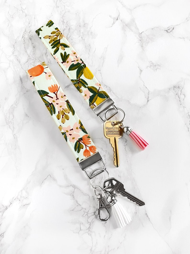 May include: Two floral print wrist keychains with silver hardware. One keychain has a pink tassel, the other a white tassel. The fabric features a light green background with orange, pink, and yellow floral designs.