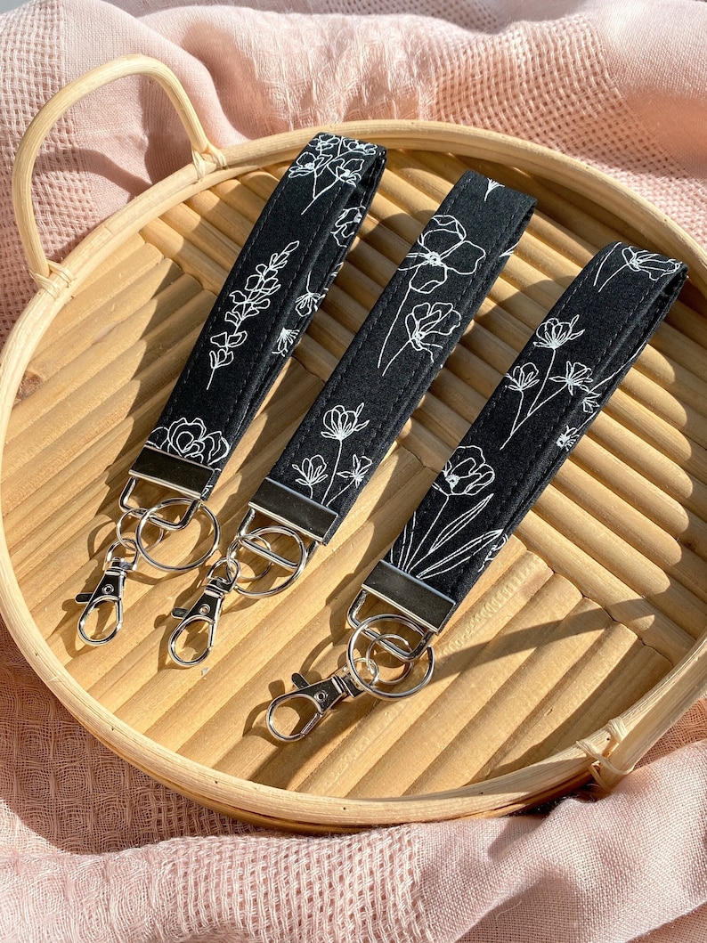 May include: Three black wrist keychains with white floral designs. Each keychain has a silver-colored clasp and ring. The keychains are arranged on a light brown, round tray, with a pink cloth in the background.