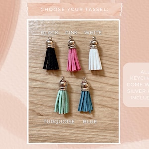 May include: A selection of keychains with different colored tassels: black, pink, white, turquoise, and blue. Each tassel has a silver-colored cap. The text "CHOOSE YOUR TASSEL" is at the top. The text "ALL KEYCHAINS COME WITH A SILVER HOOK INCLUDED!" is on the right.