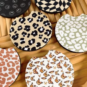 May include: A collection of car coasters with various patterns. Designs include black and white smiley faces, a black and tan checkerboard, leopard print, a sage green leopard print, a brown and white cow print, and butterflies.