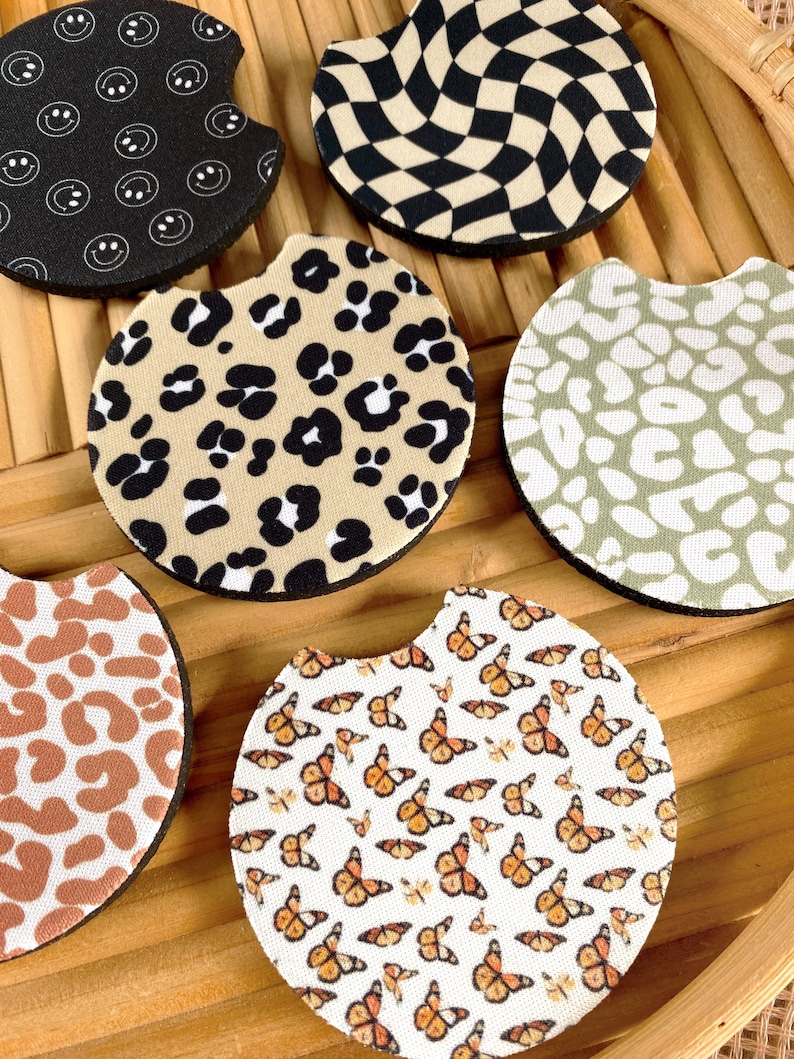 May include: A collection of car coasters with various patterns. Designs include black and white smiley faces, a black and tan checkerboard, leopard print, a sage green leopard print, a brown and white cow print, and butterflies.