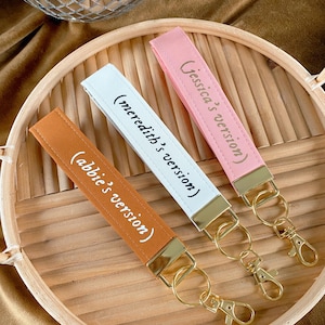May include: Three keychains with wrist straps in orange, white, and pink, each with gold-colored hardware. The straps have text that reads "(abbie's version)", "(meredith's version)", and "(Jessica's version)". The keychains are on a wooden tray.