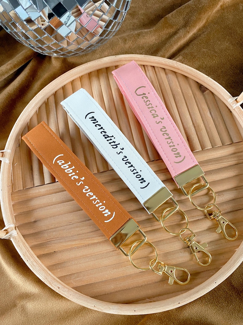 May include: Three keychains with wrist straps in orange, white, and pink, each with gold-colored hardware. The straps have text that reads "(abbie's version)", "(meredith's version)", and "(Jessica's version)". The keychains are on a wooden tray.
