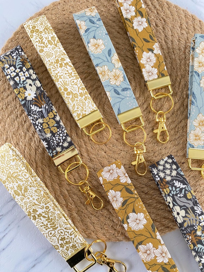 May include: A collection of floral patterned key fobs in various colors, including gold, blue, and mustard yellow. Each fob features a gold-colored metal clasp and key ring. The fabric designs showcase white flowers and foliage.