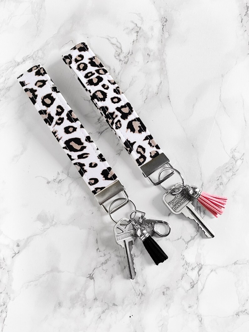May include: Two key wristlets with a white background and a black and tan leopard print pattern. Each wristlet has a silver-colored metal clasp and key ring. One wristlet has a black tassel, and the other has a pink tassel.