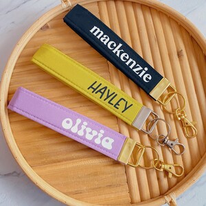 May include: Three personalized wrist keychains in navy, yellow, and lavender, each with a name in white lettering. The keychains have gold or silver-colored hardware and are displayed on a round, woven tray.