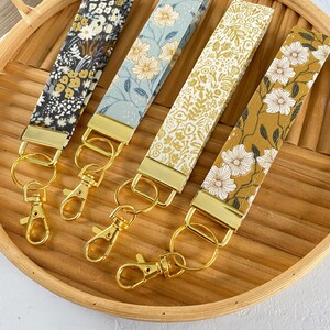 May include: Four fabric wrist keychains with floral patterns in black, blue, cream, and mustard yellow. Each keychain has a gold-colored metal clasp and ring. The keychains are displayed on a wooden tray.