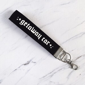 May include: Black wristlet key fob with the words "getaway car" in white gothic font. The key fob has a silver metal clasp and key ring. The background is a white marble surface.