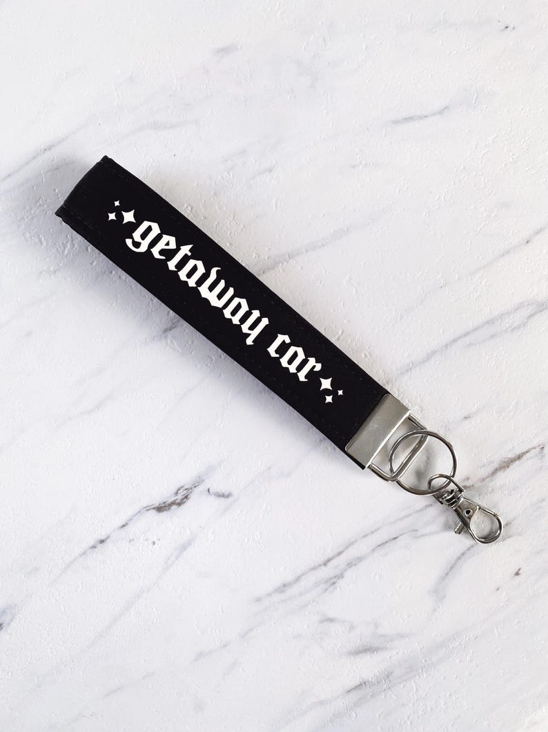 May include: Black wristlet key fob with the words "getaway car" in white gothic font. The key fob has a silver metal clasp and key ring. The background is a white marble surface.