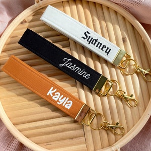 May include: Three personalized wrist keychains in white, black, and orange. Each keychain features a name embroidered in a contrasting color: "Sydney", "Jasmine", and "Kayla". Gold-colored hardware includes a clasp and a key ring. The keychains are displayed on a round, woven tray.