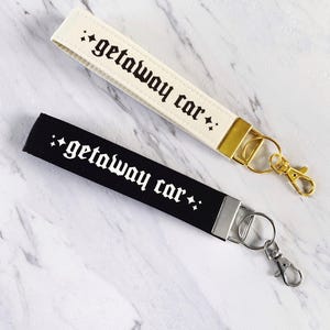 May include: Two wrist keychains, one cream and one black, each with the phrase "getaway car" in gothic font. The cream keychain has gold hardware, while the black one has silver hardware. Both have a small star design.