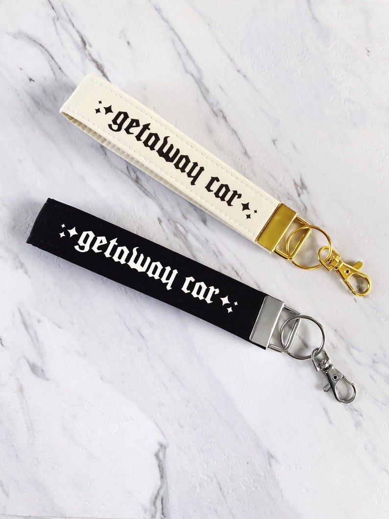 May include: Two wrist keychains, one cream and one black, each with the phrase "getaway car" in gothic font. The cream keychain has gold hardware, while the black one has silver hardware. Both have a small star design.