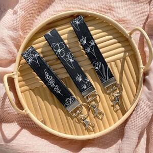 May include: Three black fabric keychains with white floral designs, each featuring a silver-tone clasp and ring. The keychains are arranged on a round, light brown woven tray, set against a soft pink background.