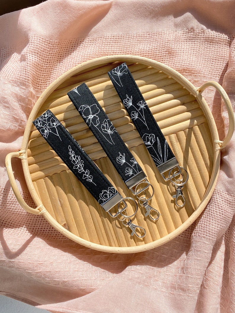 May include: Three black fabric keychains with white floral designs, each featuring a silver-tone clasp and ring. The keychains are arranged on a round, light brown woven tray, set against a soft pink background.