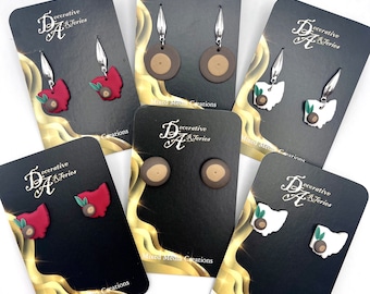 May include: Six pairs of earrings displayed on cards. The earrings are made from polymer clay and feature a variety of designs, including a brown circle, a red Ohio state outline, and a white Ohio state outline. The cards are black with gold trim and feature the text "Decorative Arts & Series" and "Mixed Media Creations".
