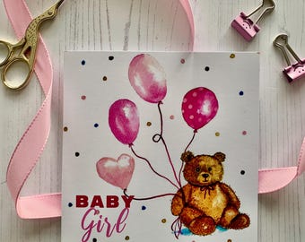 May include: A square baby girl greeting card with a watercolor illustration of a brown teddy bear holding pink balloons. The card has the words ".BABY Girl" in pink. Pink ribbon, gold scissors, and pink binder clips are in the background.
