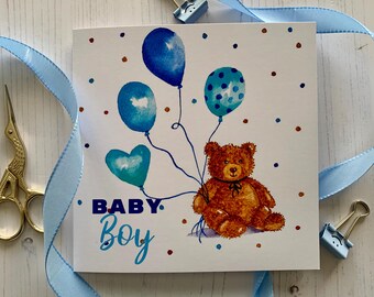 May include: A square greeting card with a watercolor illustration of a brown teddy bear holding blue balloons. The card reads "BABY BOY" in blue lettering, with blue and brown polka dots scattered around the image. A blue ribbon and gold scissors are in the background.