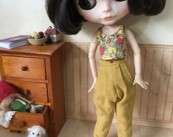 May include: A doll with black hair and freckles wearing a yellow floral tank top and mustard yellow trousers. The doll is standing on a wooden floor.