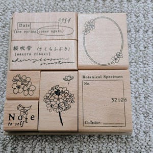 May include: A set of six light-colored wooden rubber stamps featuring various designs including a date box with the text the spring comes again, a floral oval frame, cherry blossom illustrations, the text Note to self, and a Botanical Specimen box.