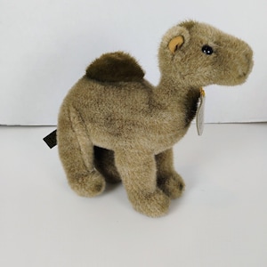 May include: A plush camel toy in shades of tan and brown. The stuffed animal has a hump, small ears, and a tag attached to its neck. The camel is standing on all fours, with a soft, textured appearance, and is set against a white background.