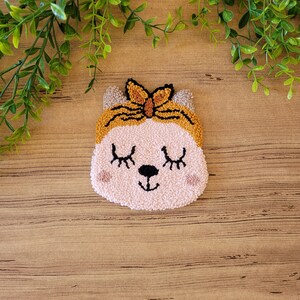Op de afbeelding: Een punch needle wandhanger met een lichtroze kattengezicht met gesloten ogen, een zwarte neus en roze wangen. De kat draagt een mosterdgele hoofdband met een strikdetail. Het getextureerde vezelkunstwerk heeft een zachte, lusvormige afwerking.