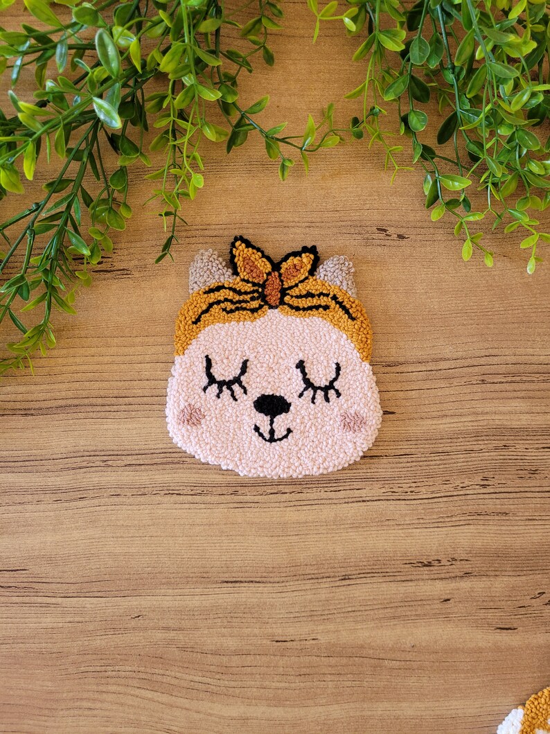 Op de afbeelding: Een punch needle wandhanger met een lichtroze kattengezicht met gesloten ogen, een zwarte neus en roze wangen. De kat draagt een mosterdgele hoofdband met een strikdetail. Het getextureerde vezelkunstwerk heeft een zachte, lusvormige afwerking.