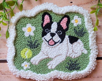 May include: A rectangular punch needle wall hanging featuring a black and white French Bulldog lying on a green background. The dog has pink inner ears and a pink tongue. The scene includes white flowers with yellow centers and a yellow tennis ball.