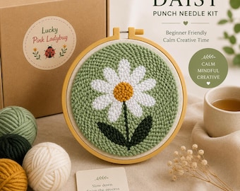 May include: A punch needle kit featuring a daisy design in a wooden hoop, a brown cardboard box labeled Lucky Pink Ladybug, four yarn balls in green, black, white, and yellow, and a card with the text Slow down Enjoy the process You've got this.
